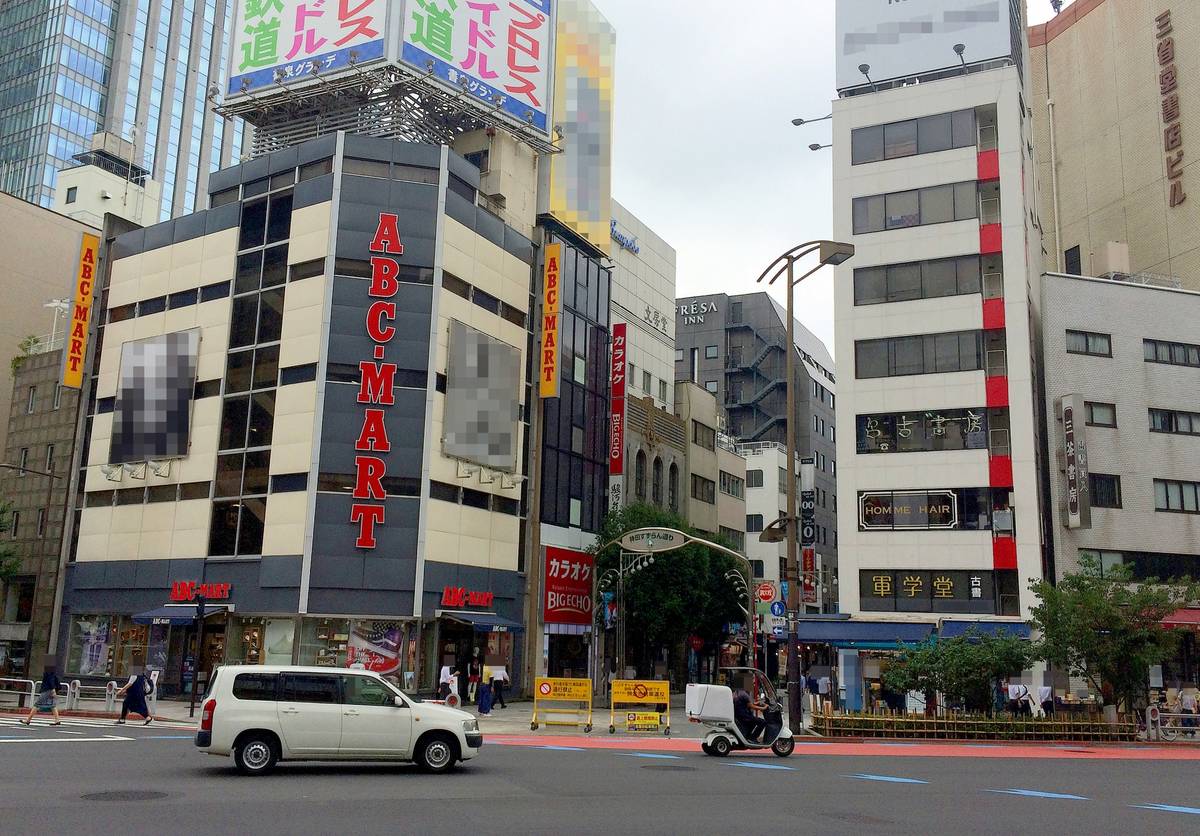 A city street with an ABC-MART building on a corner, a white van, and a delivery scooter.