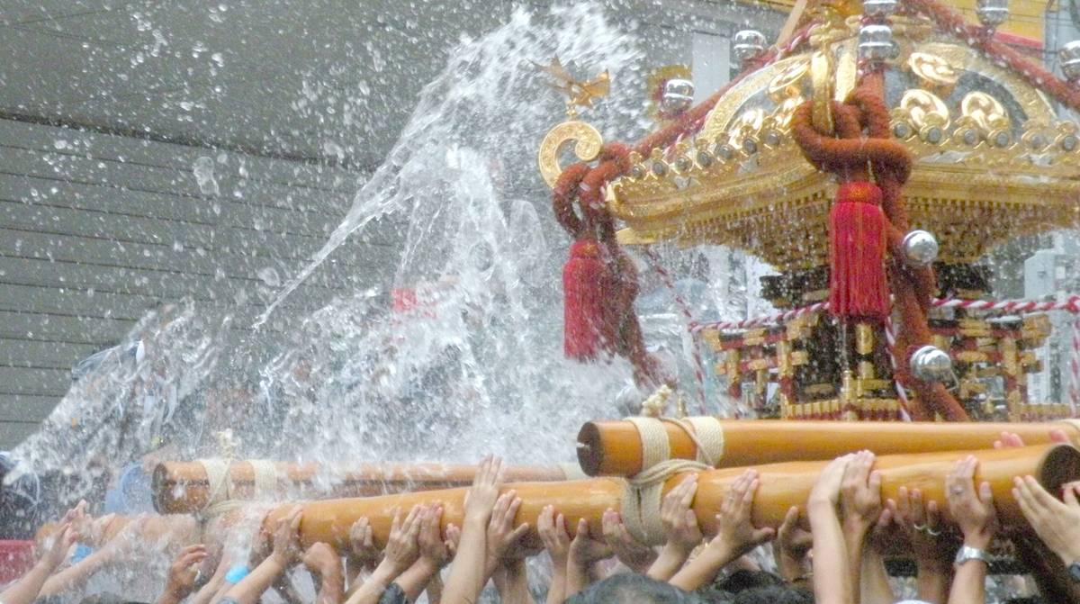 A golden, ornate mikoshi is carried by many hands as water splashes all over it during a festival.