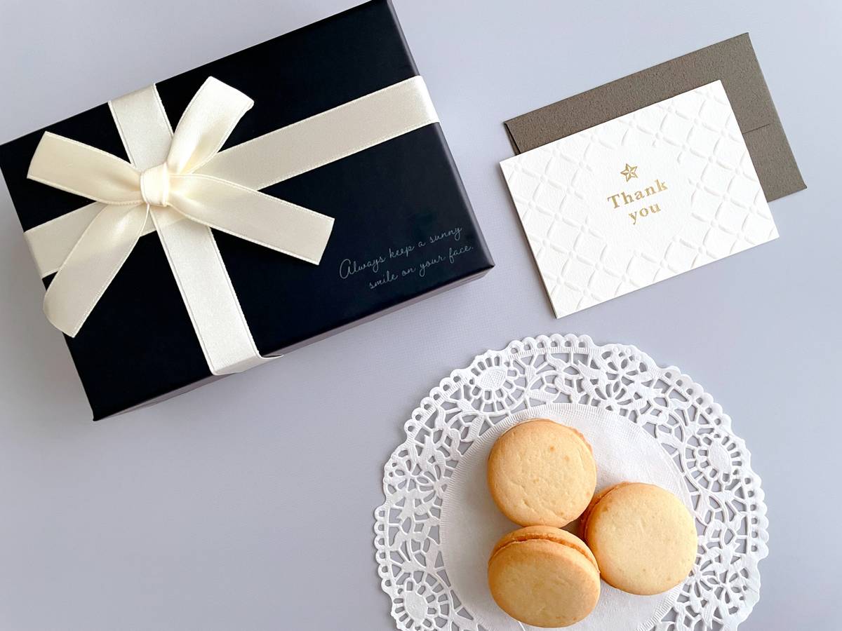 A black gift box with a cream ribbon, a 'Thank you' card, and three macarons on a lace doily.