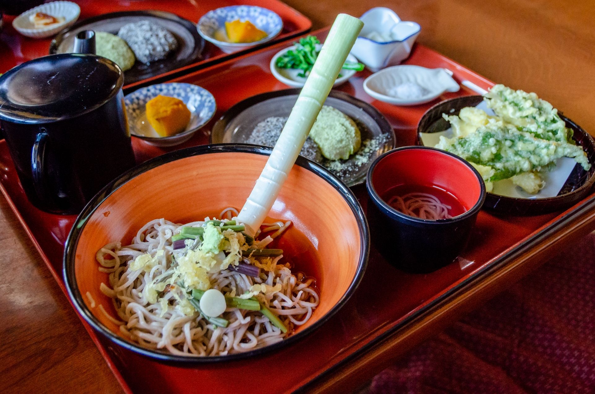 A Japanese soba noodle meal set on a red tray, featuring a large bowl of noodles with a prominent green leek