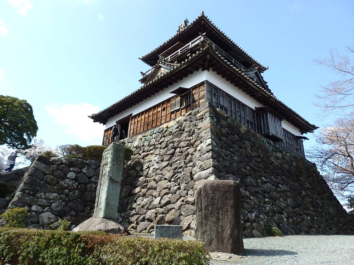A multi-story Japanese castle with dark tiled roofs stands on a tall stone foundation under a clear blue sky.