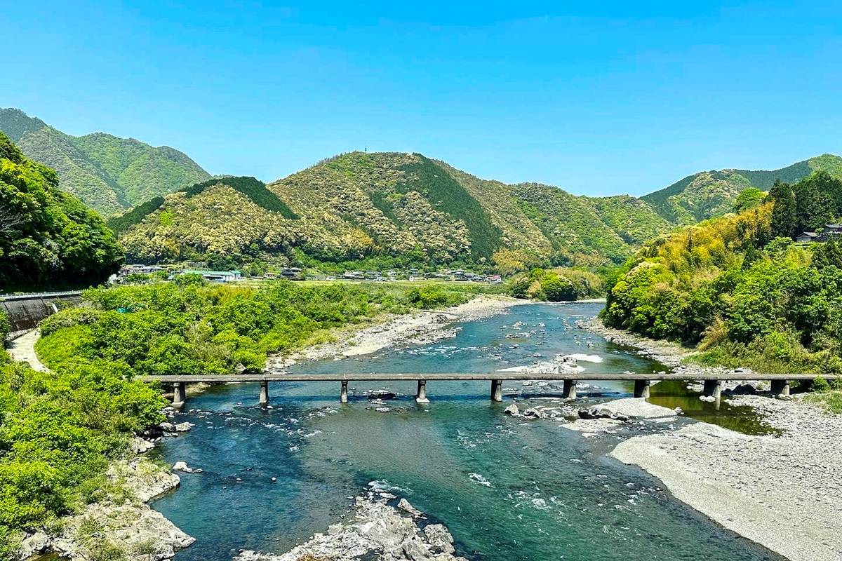 A submersible bridge crosses a wide river winding through a valley of green mountains under a clear blue sky.