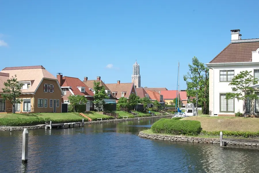 European-style houses line a canal with a sailboat and a tall church tower under a clear blue sky.