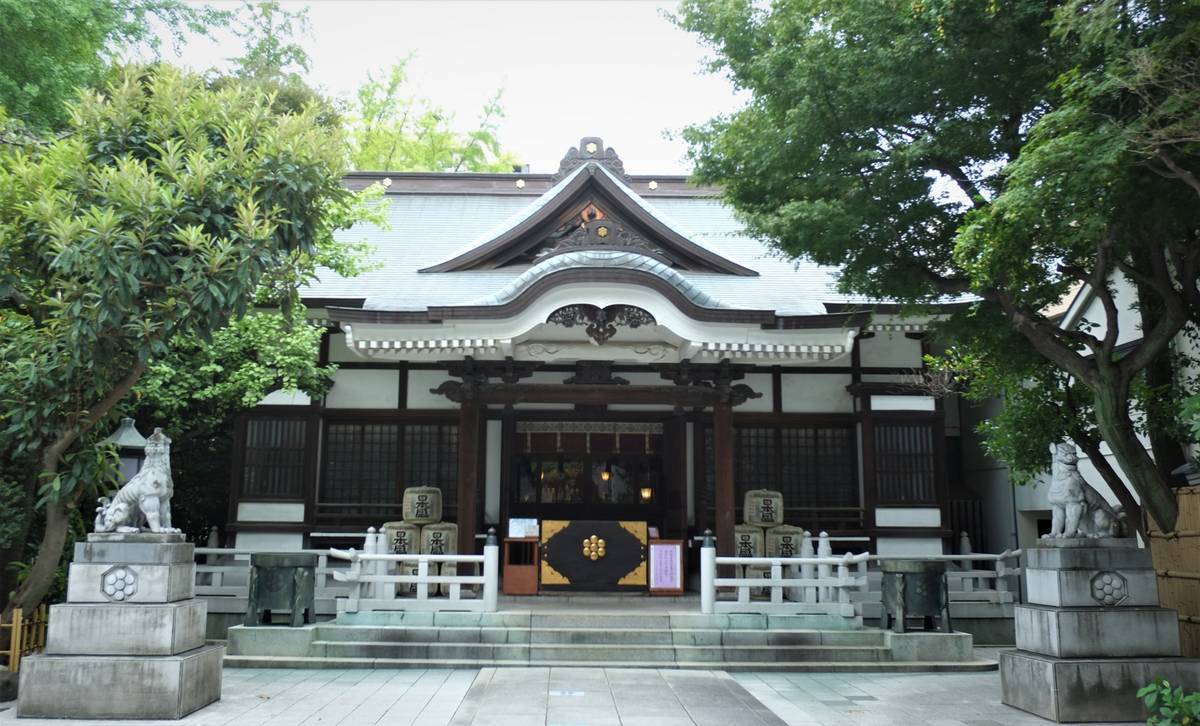A traditional Japanese shrine with a curved roof, white walls, and two stone guardian dog statues, framed by green trees.