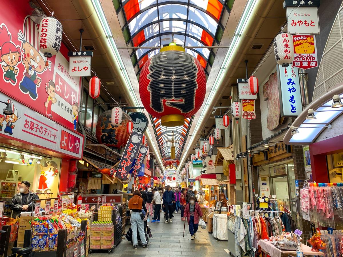 A bustling Japanese shopping arcade with people, shops, and hanging lanterns under a colorful arched roof.