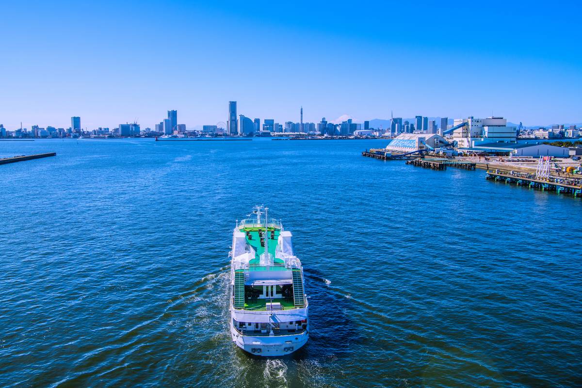 A white and green ferry sails on a bay towards a city skyline and port, with distant Mount Fuji under a clear blue sky.