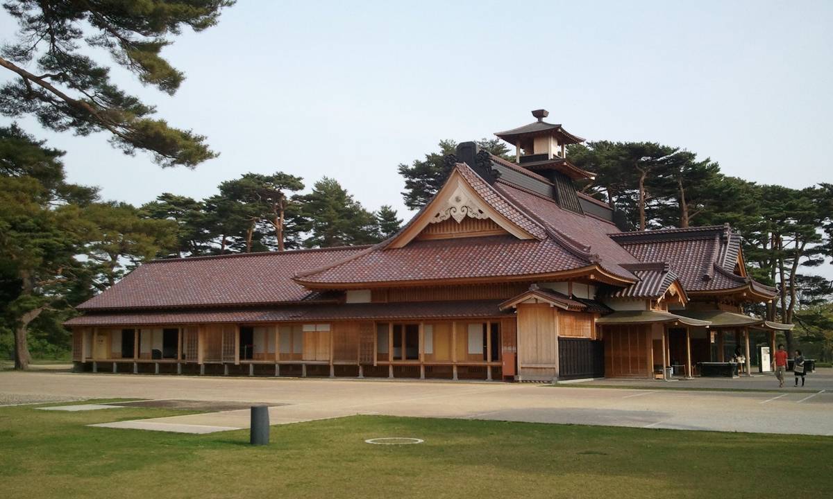 A traditional light-colored wooden Japanese building with a reddish-brown tiled roof, surrounded by pine trees.