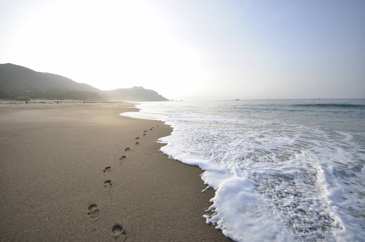 Footprints in sand beside foamy ocean waves on a bright beach with distant hills.