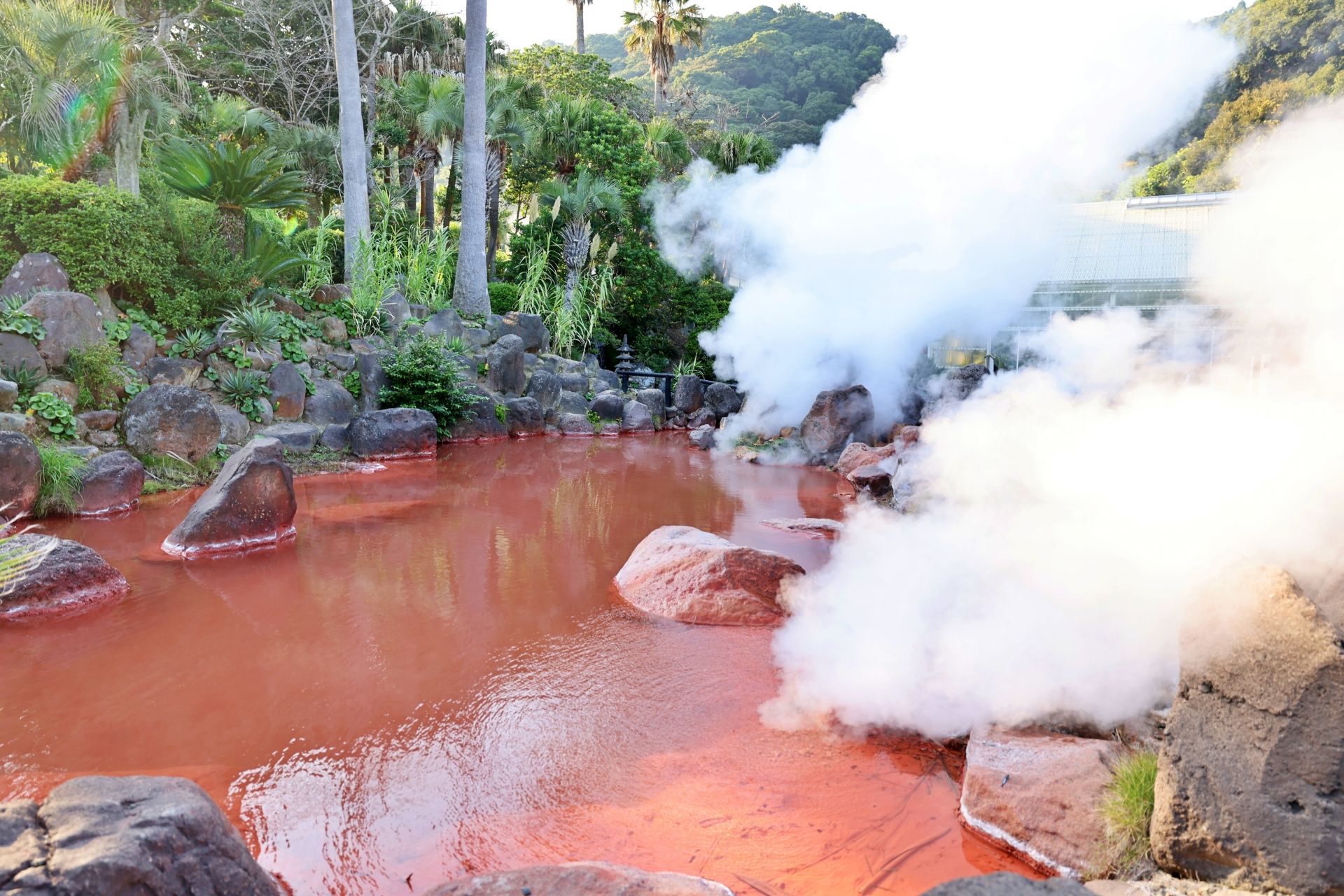 A steaming red-orange hot spring pool surrounded by rocks and lush tropical greenery.