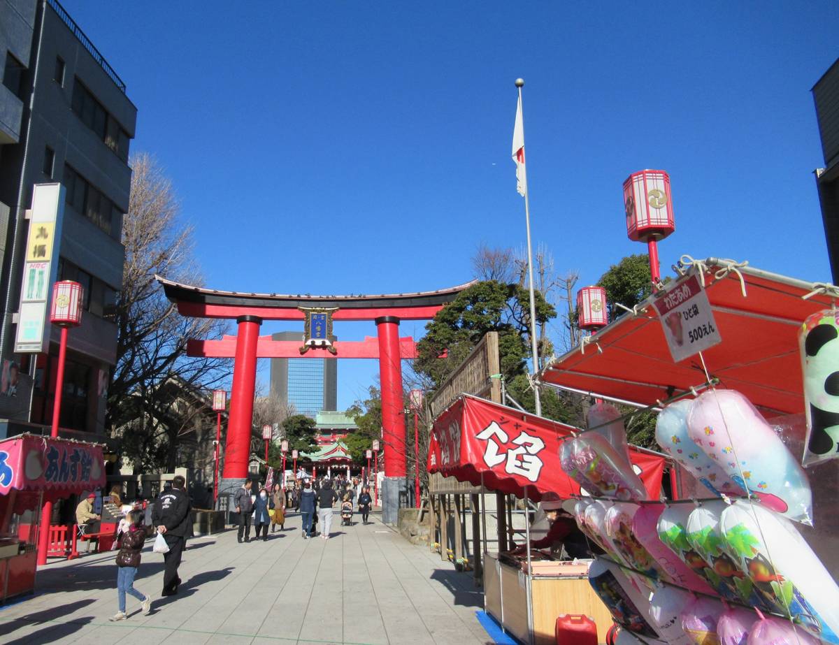A bustling street scene in Japan with a large red torii gate, festive stalls, people walking, and a Japanese flag flying under a clear blue sky.
