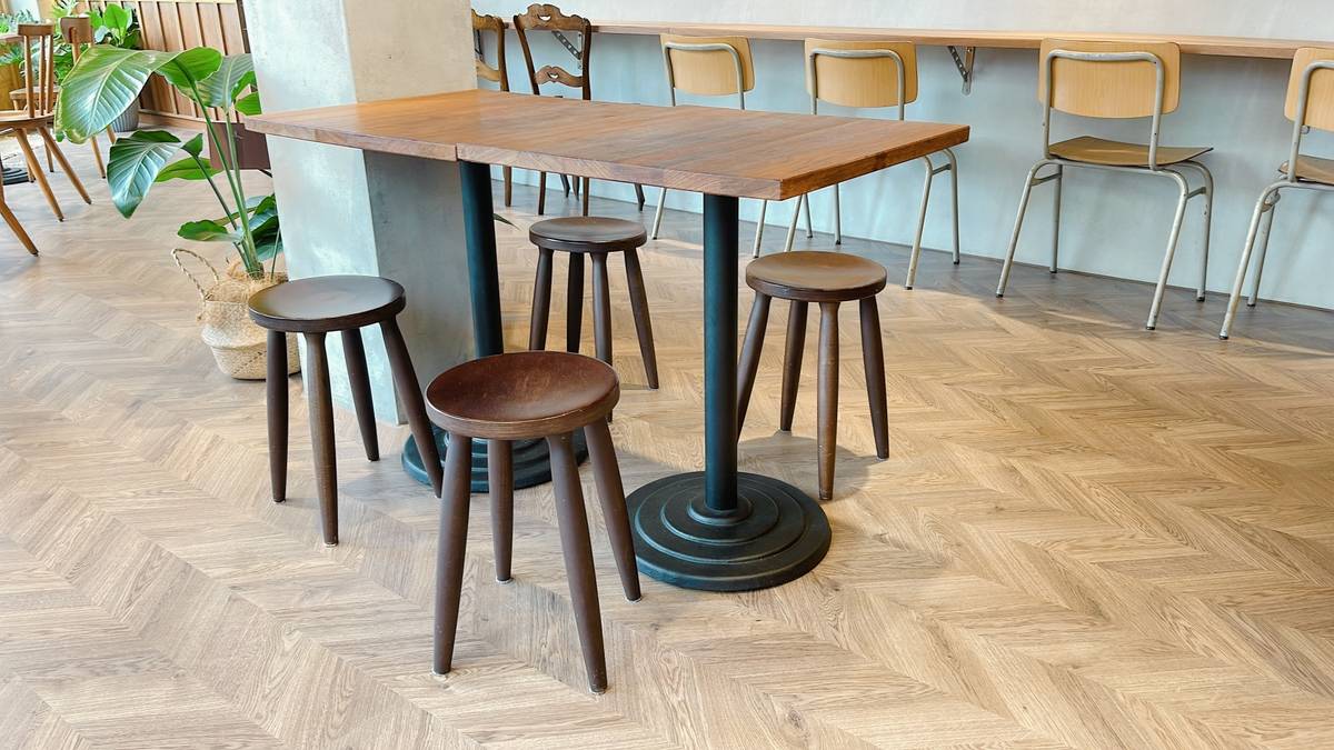 A cafe interior with a long wooden table, four dark stools, and a herringbone floor.