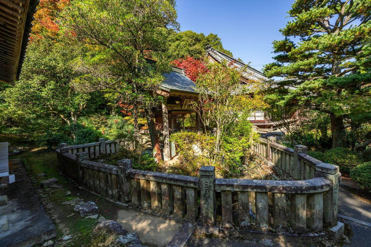 A stone fence encloses a Japanese garden with traditional buildings and trees with autumn foliage.