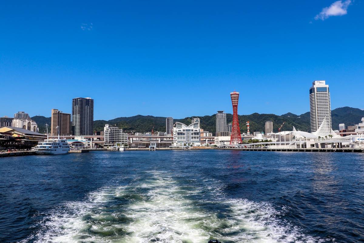 Kobe, Japan, cityscape from a bay, featuring the red Port Tower, waterfront buildings, mountains, and a boat wake.