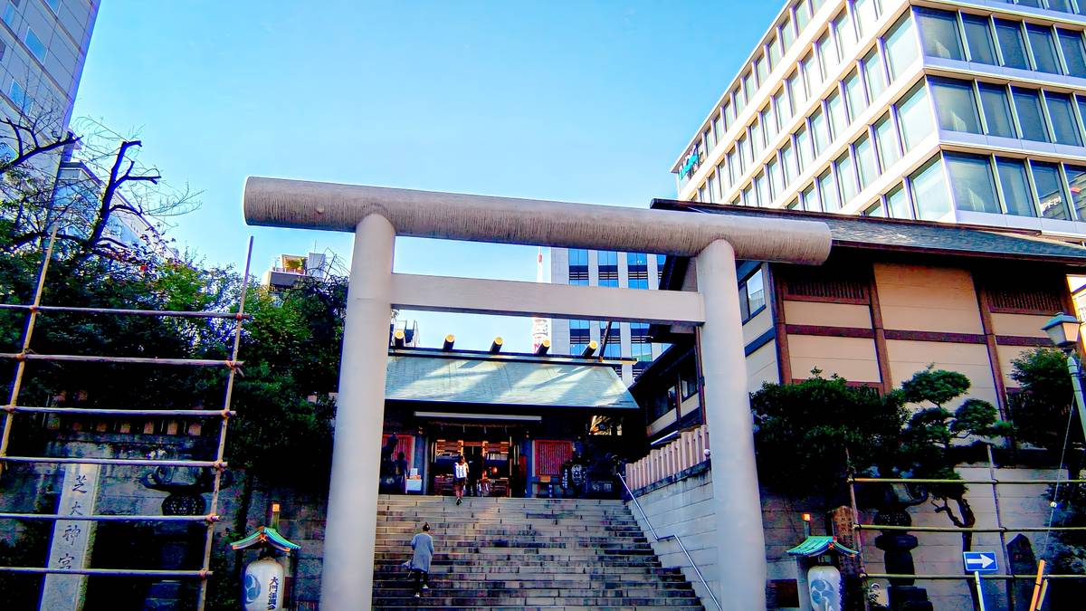 A grey torii gate at the base of stairs leading to a traditional Japanese shrine, with modern buildings in the background.