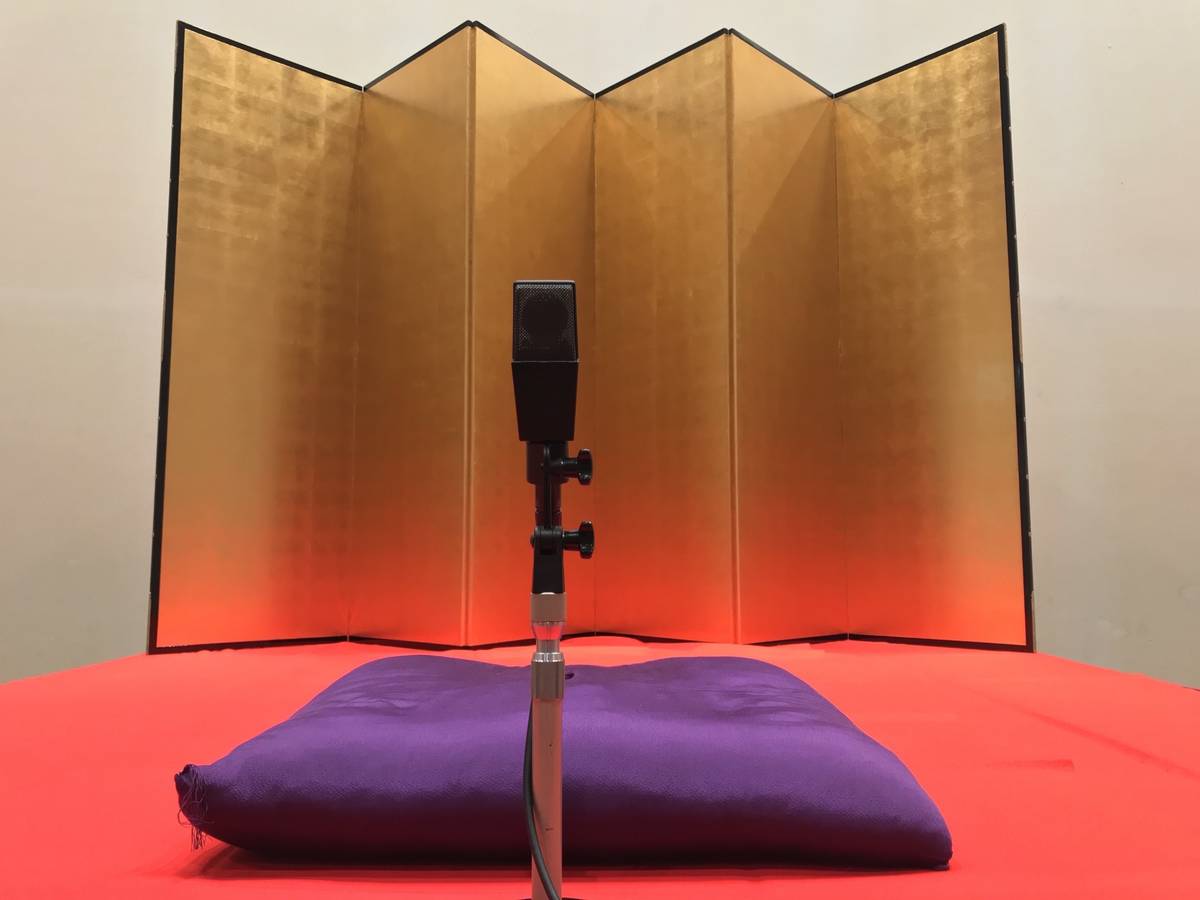 A black microphone stands before a purple cushion on a red surface, with a golden folding screen in the background.