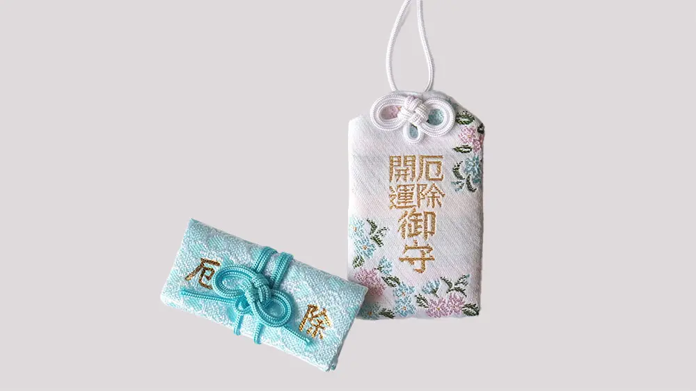 Two Japanese charms: a white rectangular hanging charm with floral embroidery and text