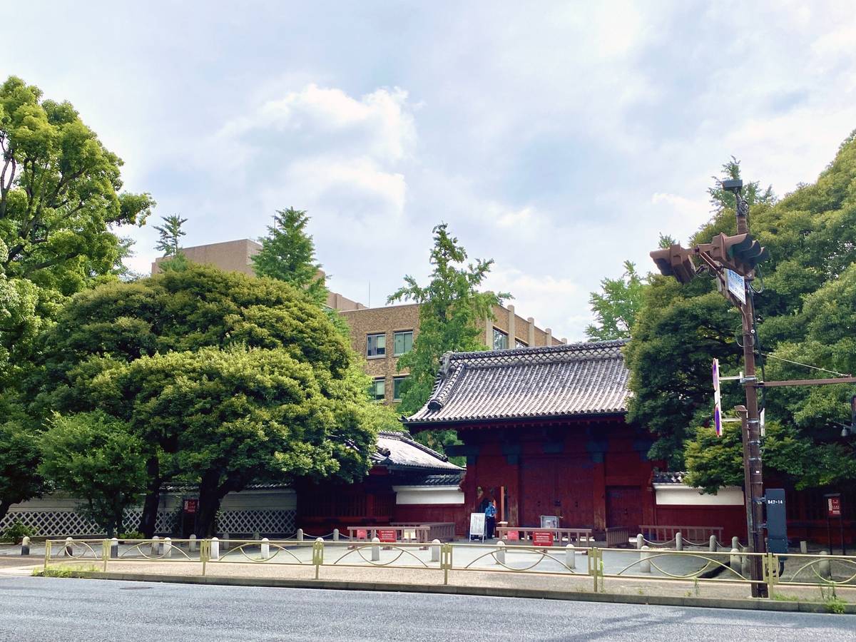 Red traditional Japanese gate with a tiled roof, surrounded by green trees, next to a street.