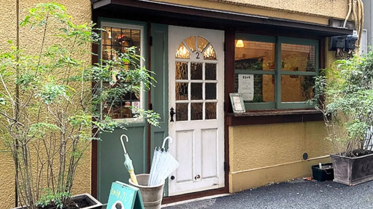 Storefront with a yellow wall, white arched door, green windows, potted plants, and umbrellas in a bucket.