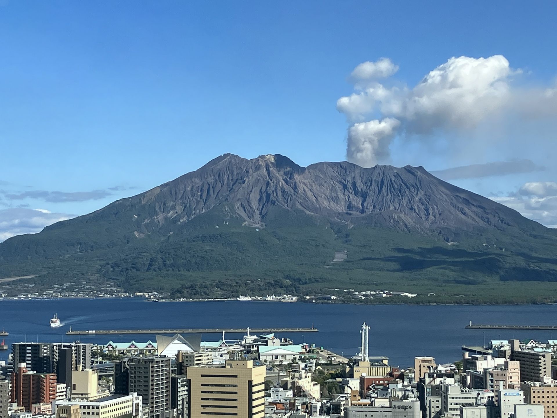 A volcano erupts smoke against a blue sky, overlooking a bay and city.