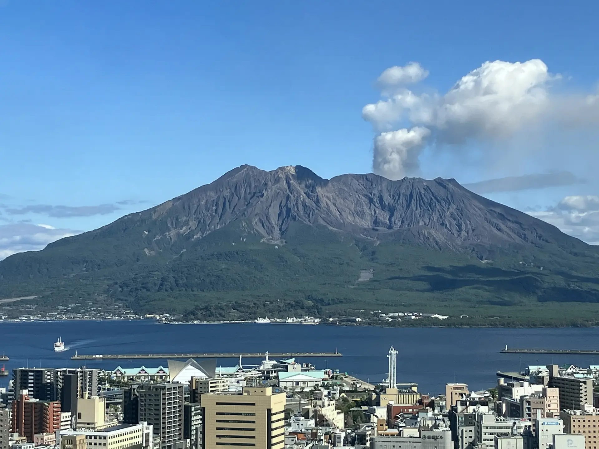 A volcano erupts smoke against a blue sky, overlooking a bay and city.