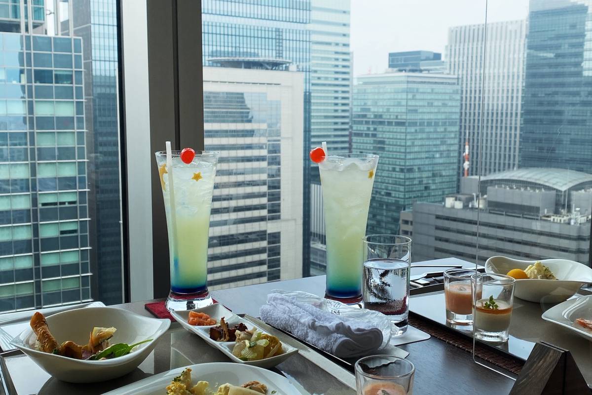 Two colorful cocktails and small dishes on a table overlooking a city skyline.