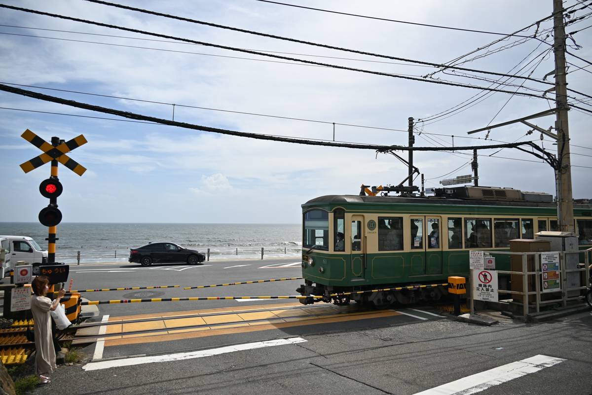 A green tram crosses a coastal railroad intersection with a woman waiting, and the ocean visible in the background.