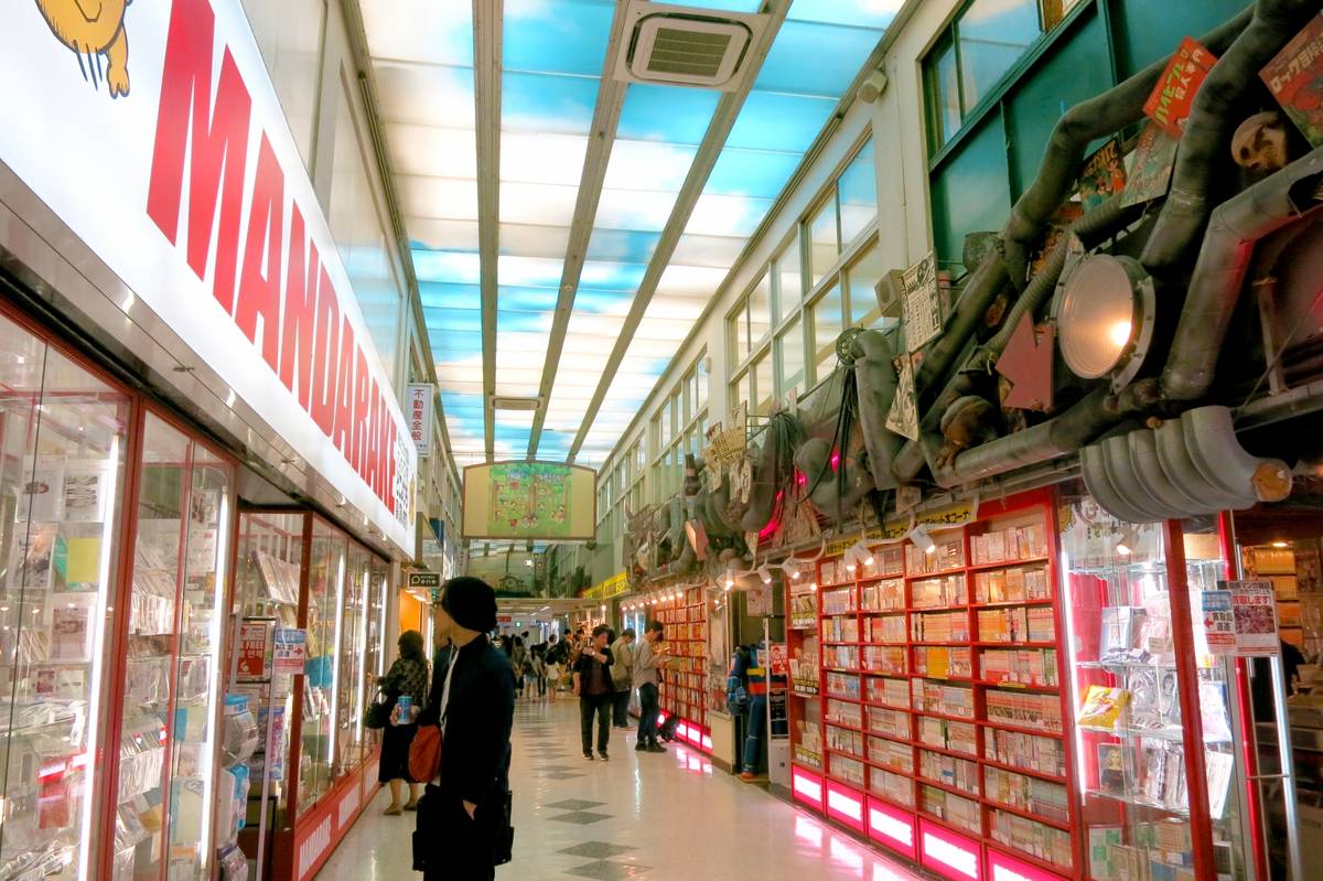 A long aisle in the Mandarake store with various merchandise, a blue sky ceiling, and industrial-style decor overhead.