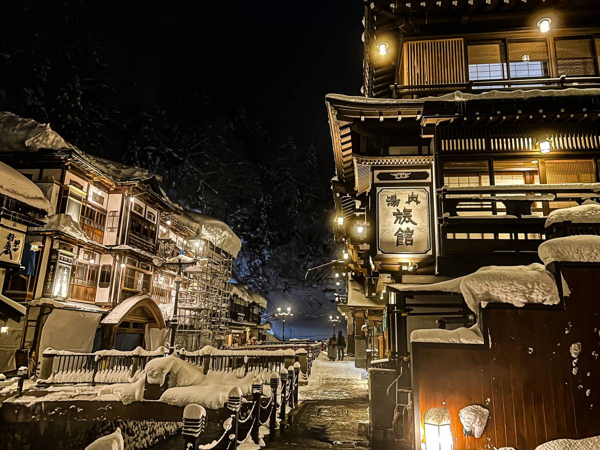A snow-covered traditional Japanese village street illuminated at night with warm lights.