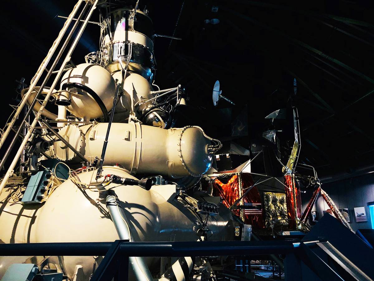 A large, light-colored spacecraft module sits beside a gold-foil lunar lander in a dimly lit museum.