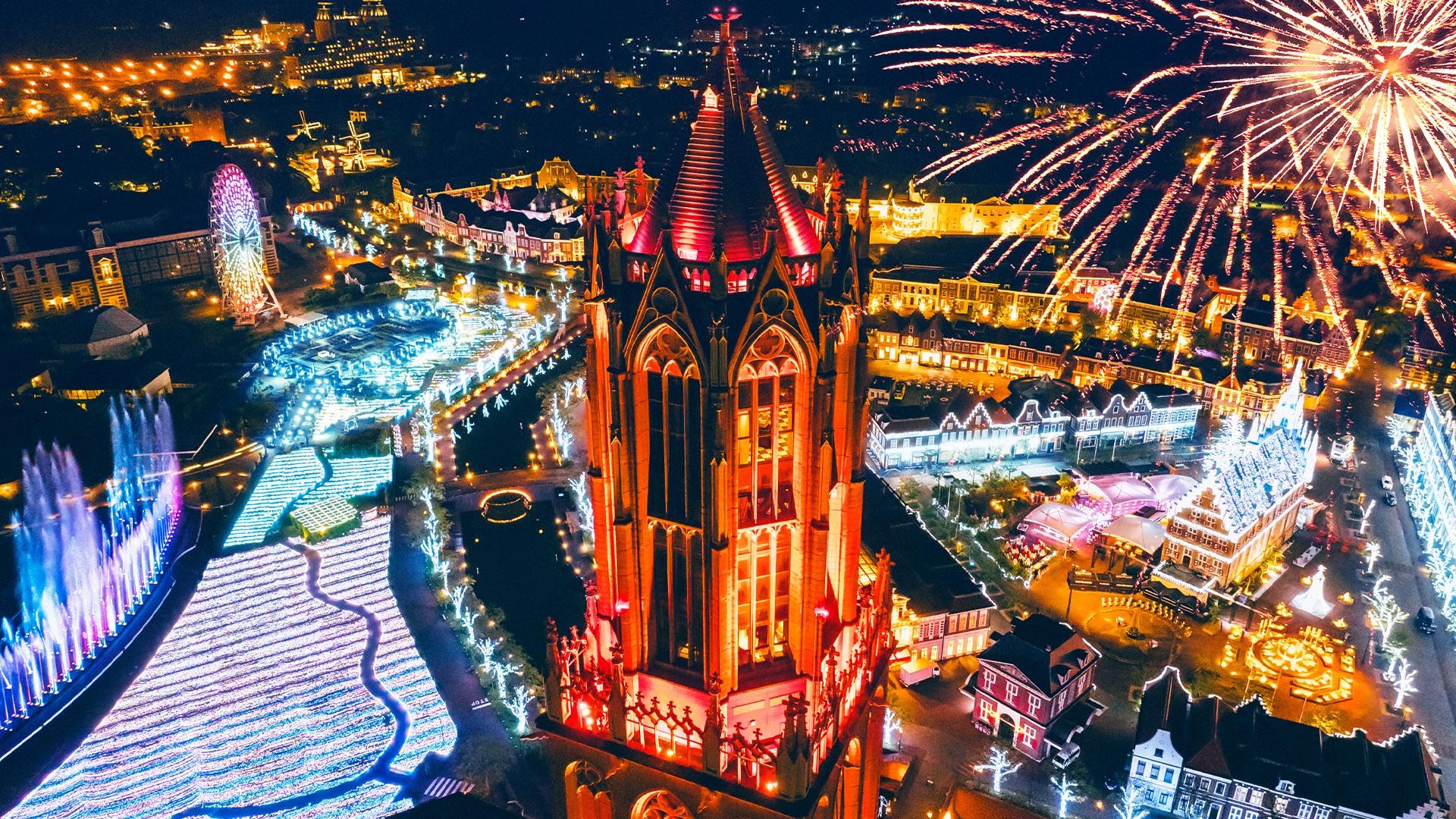 Aerial night view of a theme park featuring a red-lit gothic tower, elaborate light displays, a Ferris wheel, and fireworks.