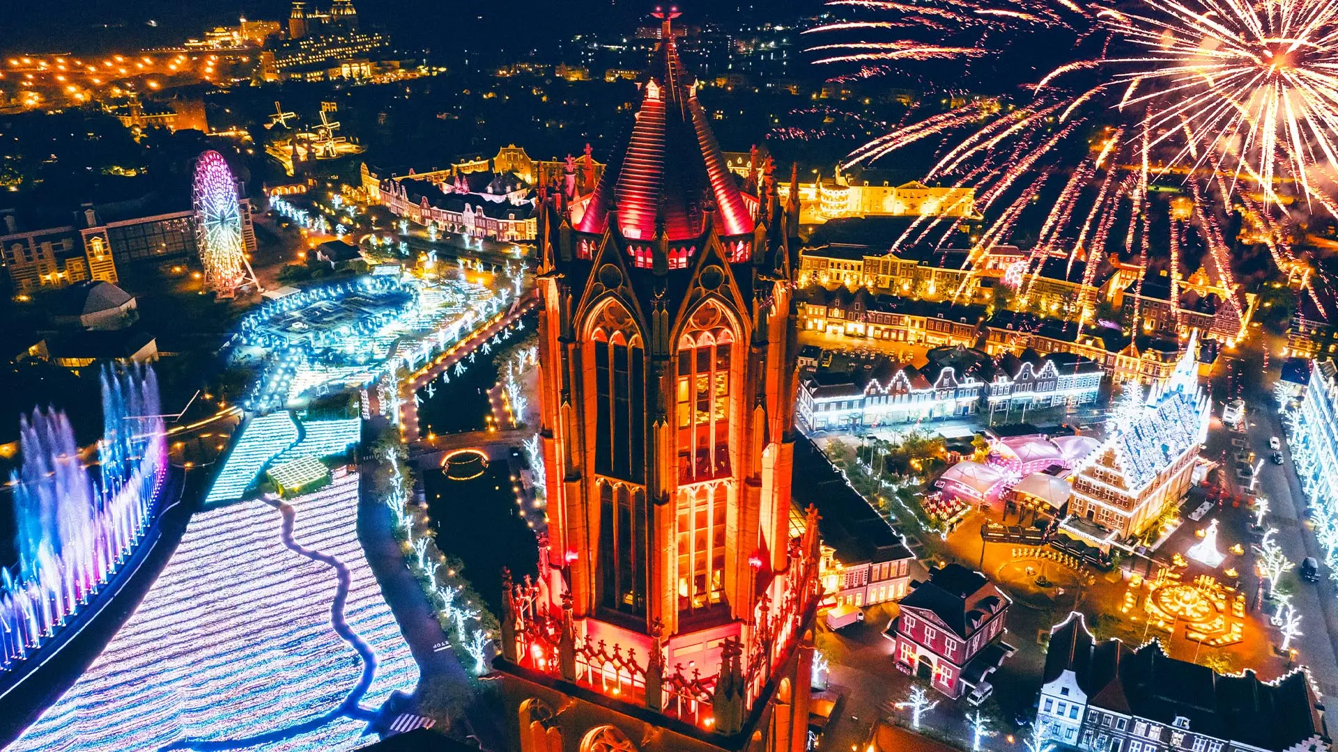 Aerial night view of a theme park featuring a red-lit gothic tower, elaborate light displays, a Ferris wheel, and fireworks.