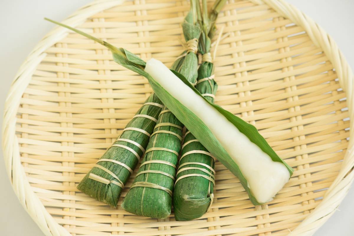 Four leaf-wrapped rice cakes on a bamboo tray, one revealing a white rice cake.