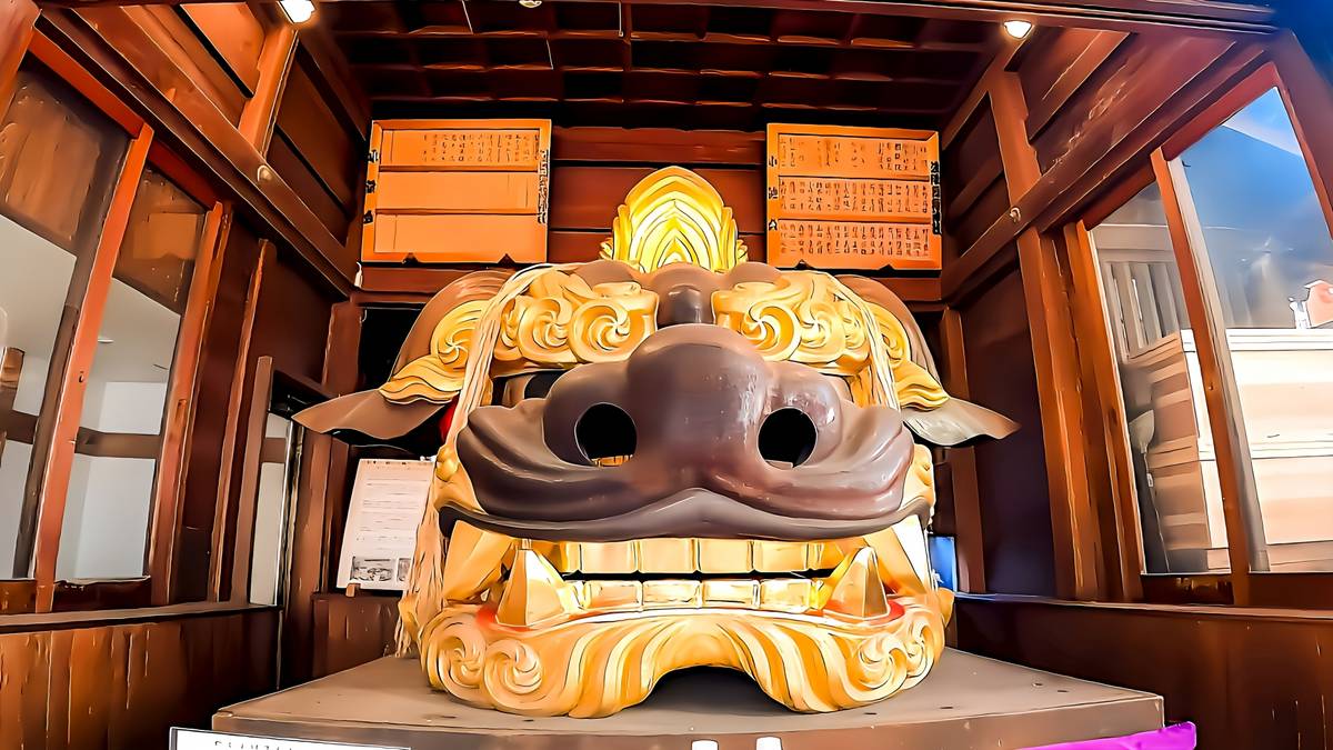 A large, ornate golden and dark brown Japanese lion-dog mask displayed in a wooden room.