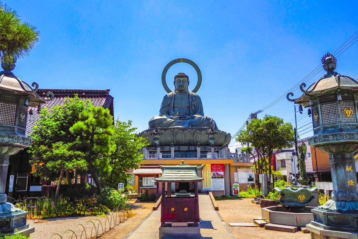 A large bronze seated Buddha statue with a halo behind its head, in a temple courtyard flanked by traditional lanterns