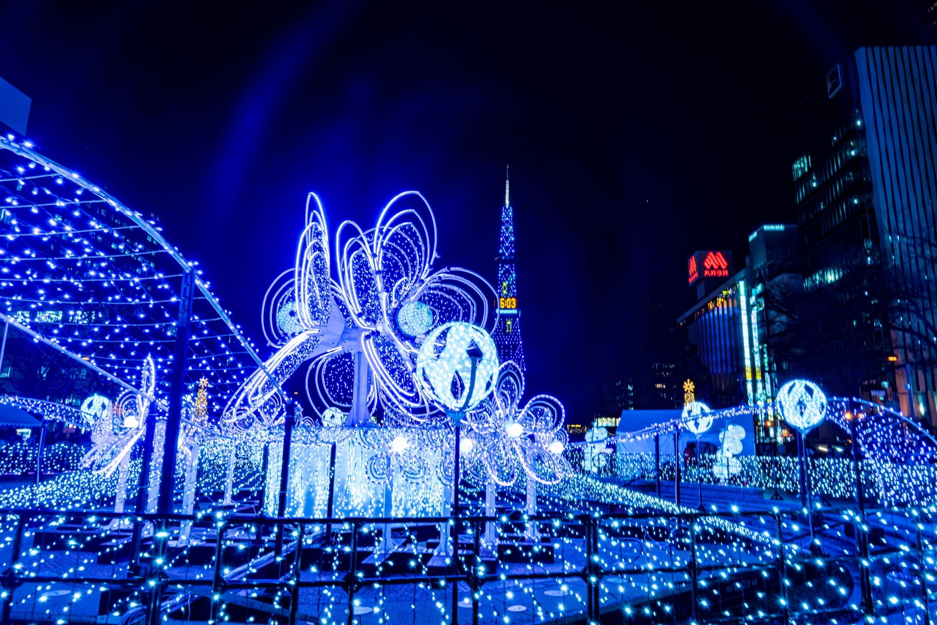 A public space at night adorned with elaborate blue and white light displays and an illuminated tower.