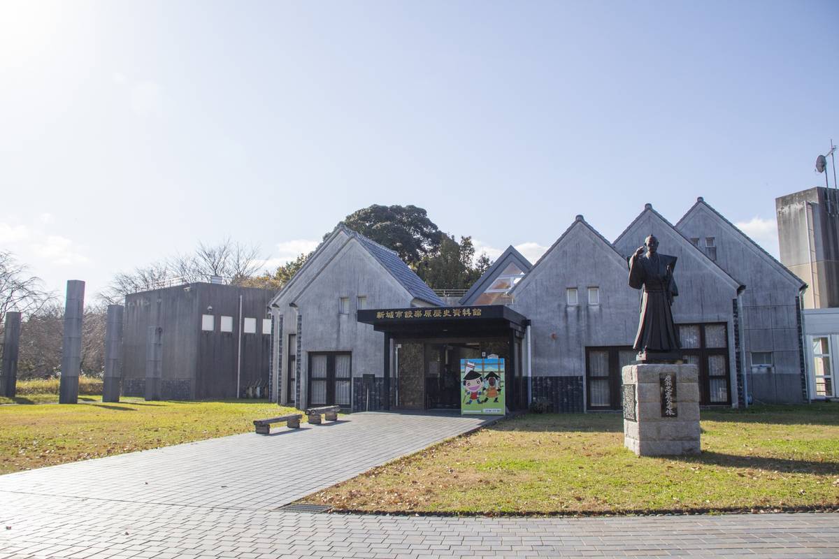 The Shinshiro City Historical Museum, a grey building with multiple gabled roofs, a statue of a robed figure, and a paved entrance path.