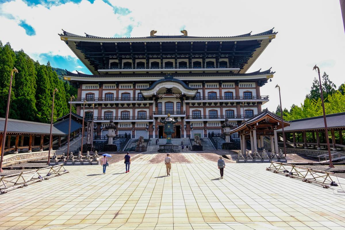 A grand Japanese temple with a dark, multi-tiered roof stands before a paved courtyard with people walking, backed by green mountains.