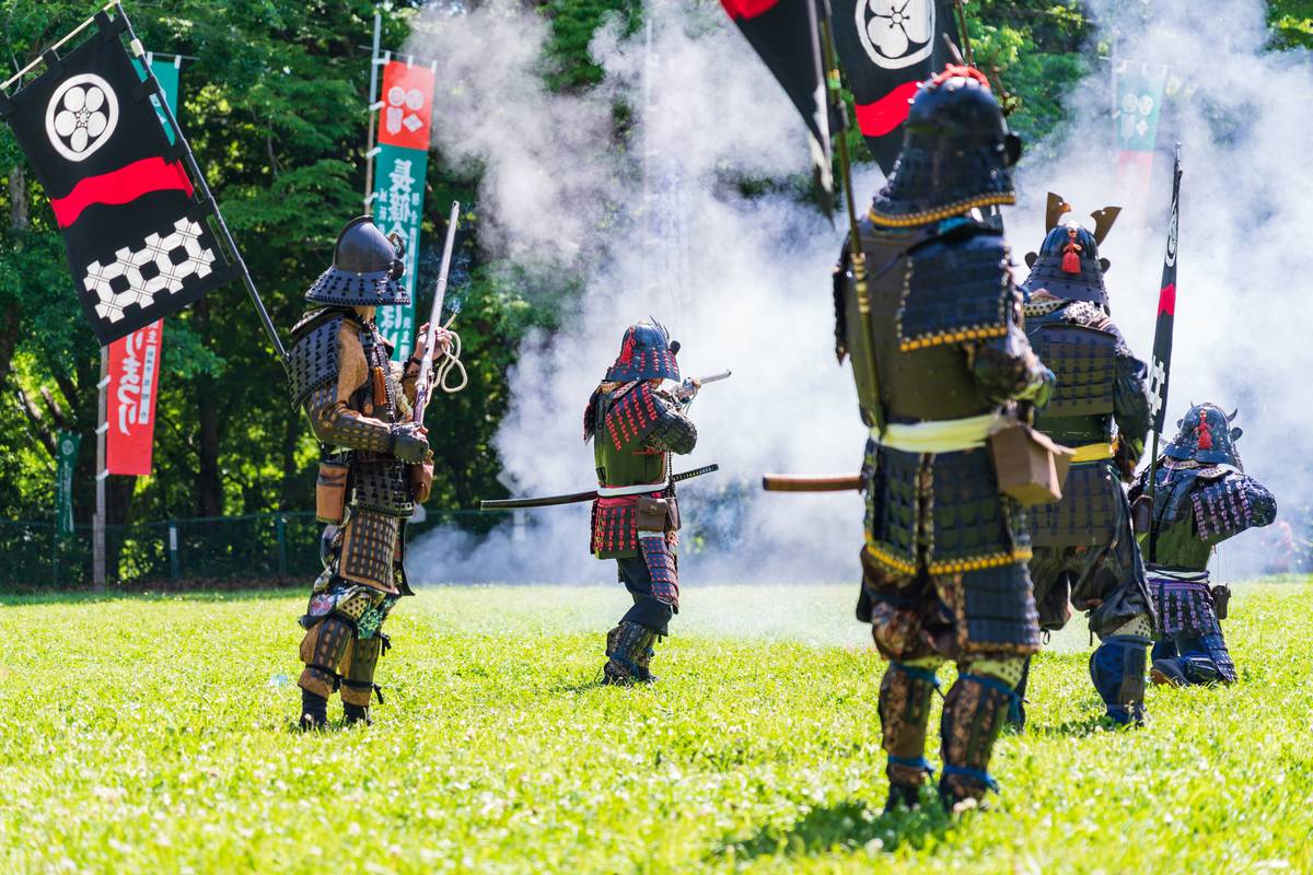 Samurai in armor fire muskets in a grassy field, with smoke and flags around them.