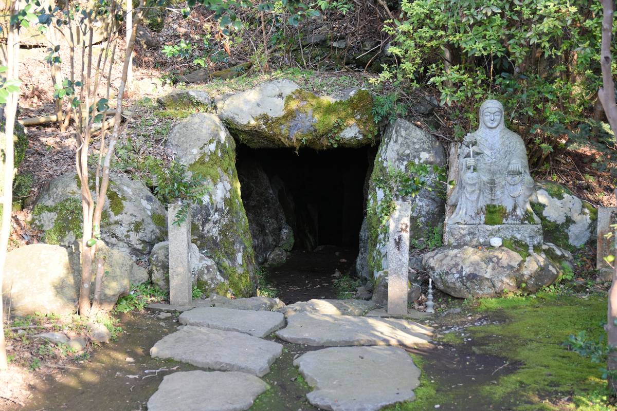 A stone path leads to a dark cave entrance formed by mossy rocks, with a large stone statue of a seated, robed figure holding a staff to its right.