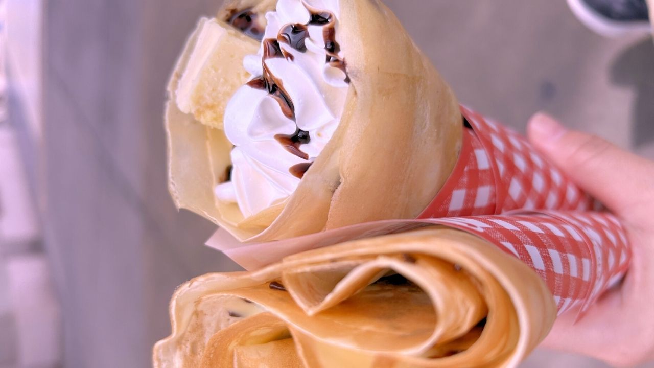 A hand holds two crepes, one topped with whipped cream, chocolate drizzle