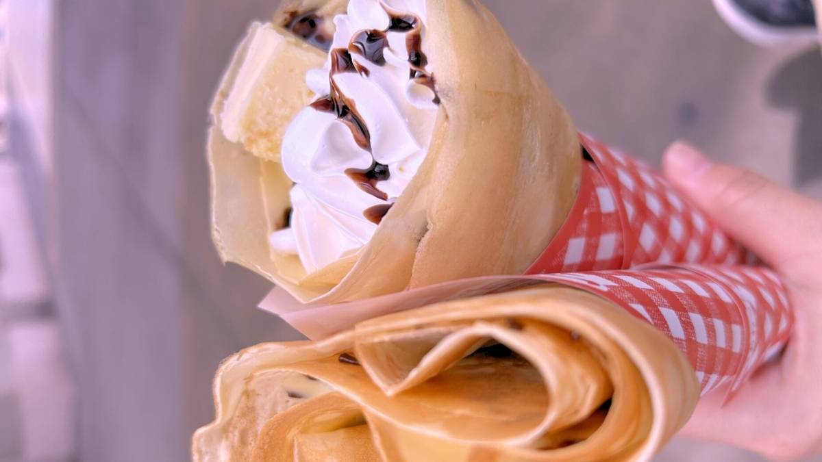 A hand holds two crepes, one topped with whipped cream, chocolate drizzle