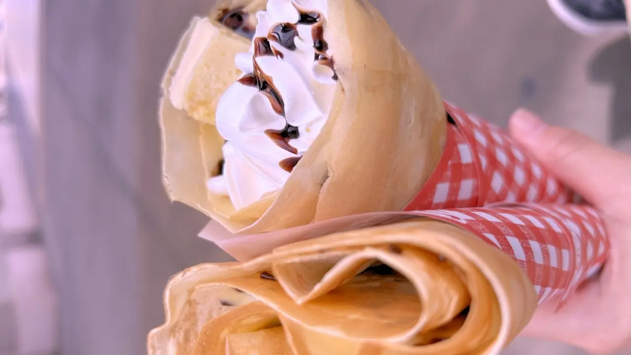 A hand holds two crepes, one topped with whipped cream, chocolate drizzle