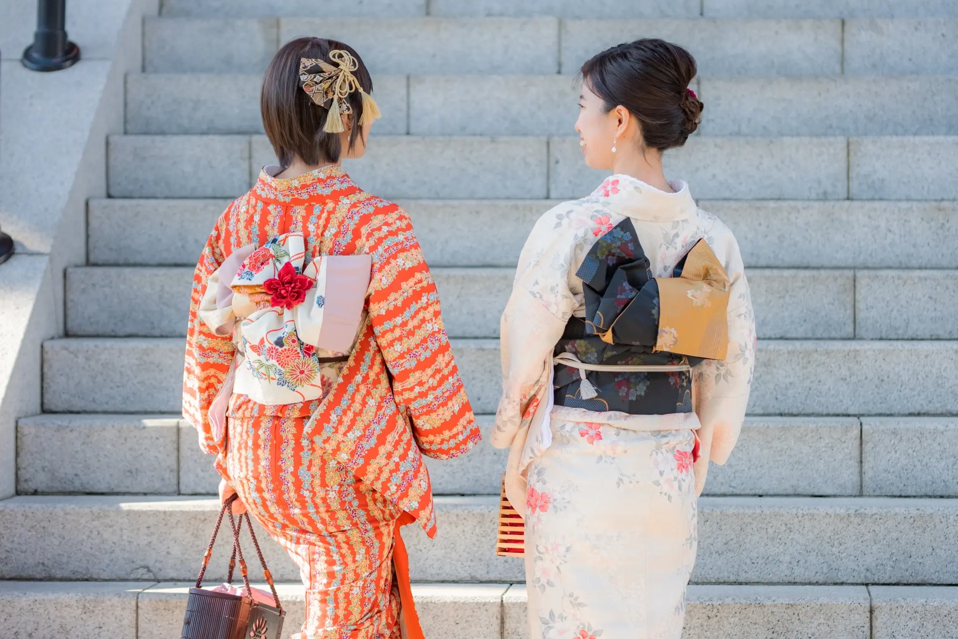 Two women in colorful kimonos stand with their backs to the viewer on stone steps.