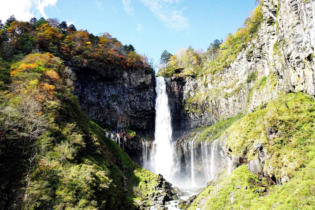 A tall waterfall cascades between rocky cliffs, flanked by trees with autumn colors.