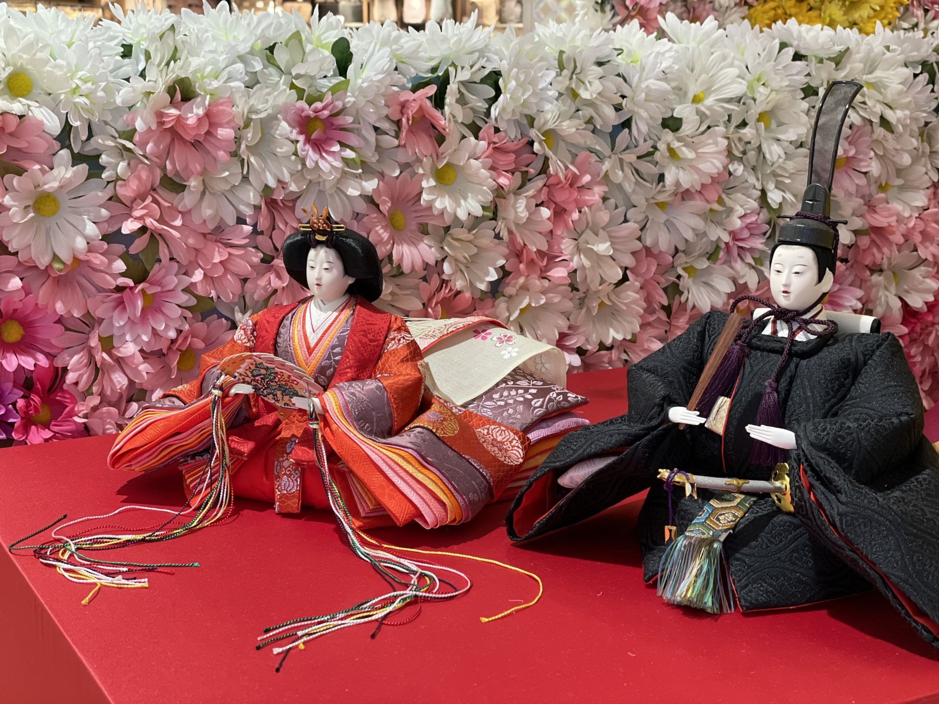 Two Hina dolls, an empress in red and an emperor in black, on a red platform in front of a wall of pink and white flowers.