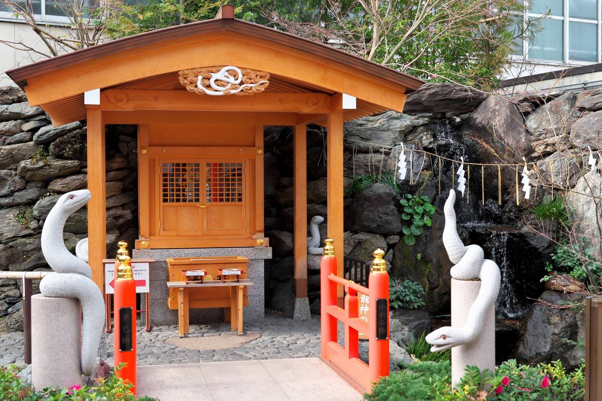 A small wooden Japanese shrine with white snake statues, a red gate, and a rock waterfall.