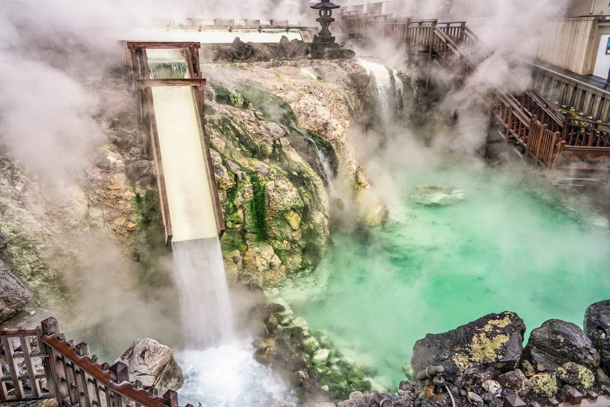 Steaming turquoise hot spring with a wooden water flume in a rocky landscape.