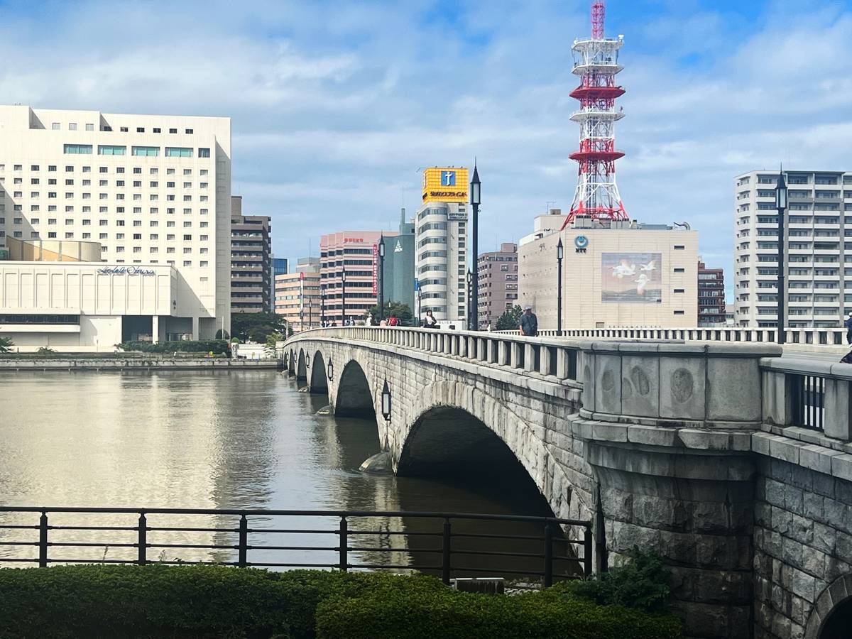 A cityscape with a stone arch bridge over water, a large white building on the left, and a red-and-white broadcast tower on the right.