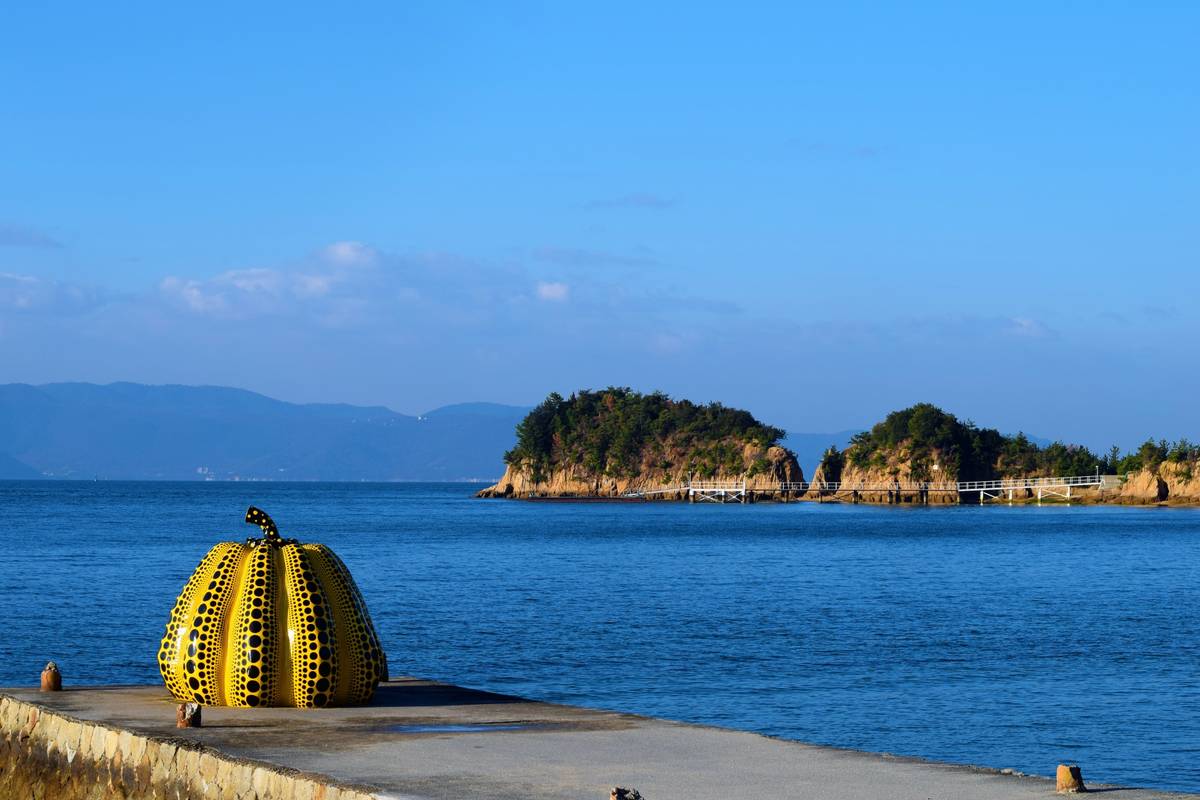 Yayoi Kusama's yellow pumpkin sculpture with black polka dots on a pier overlooking the sea and distant islands.