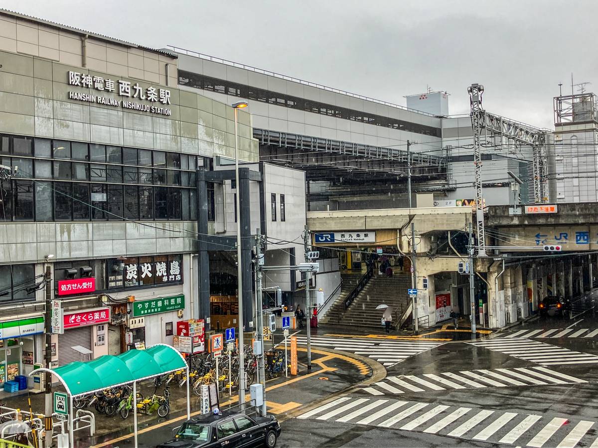 A rainy street scene in Japan showing Hanshin Railway Nishikujo Station, a JR station entrance, and a complex intersection.